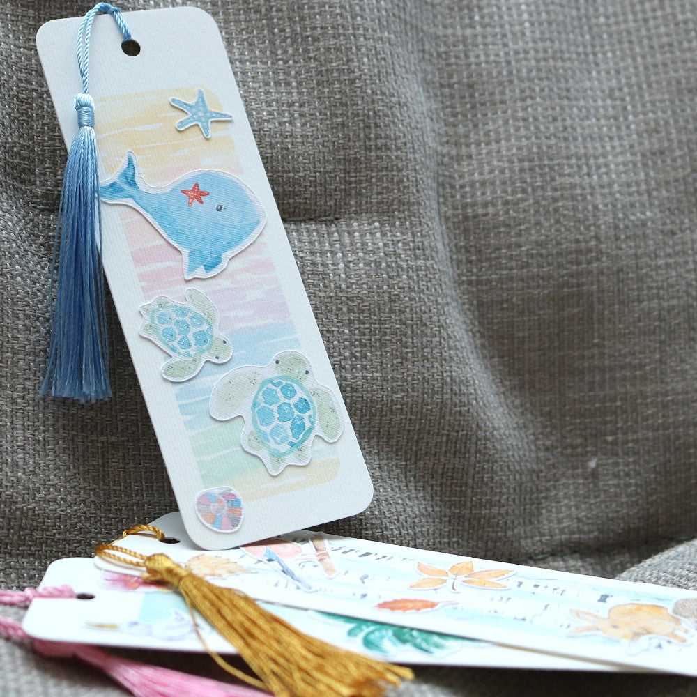 Drei Lesezeichen, eines aufgestellt mit Walen und Schildkröten, die anderen beiden darunter liegend; mit Quasten Lesezeichenbändchen