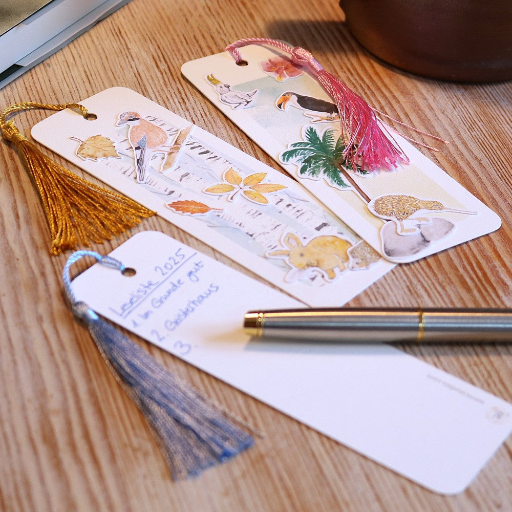 3 Lesezeichen mit Aquarellmotiven auf einem Tisch liegend, eines umgedreht und beschrieben mit daneben liegendem Stift. 